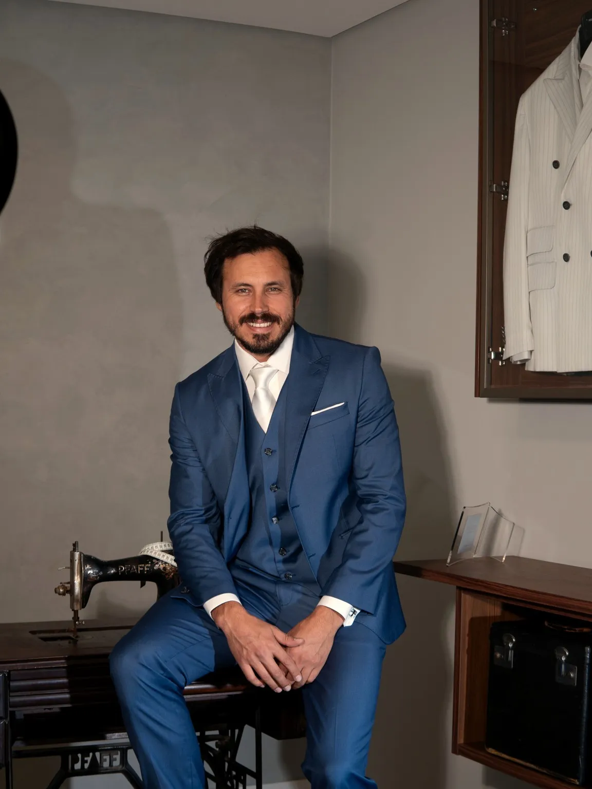 Terno azul royal com colete, sentado na mesa de costura, sorrindo