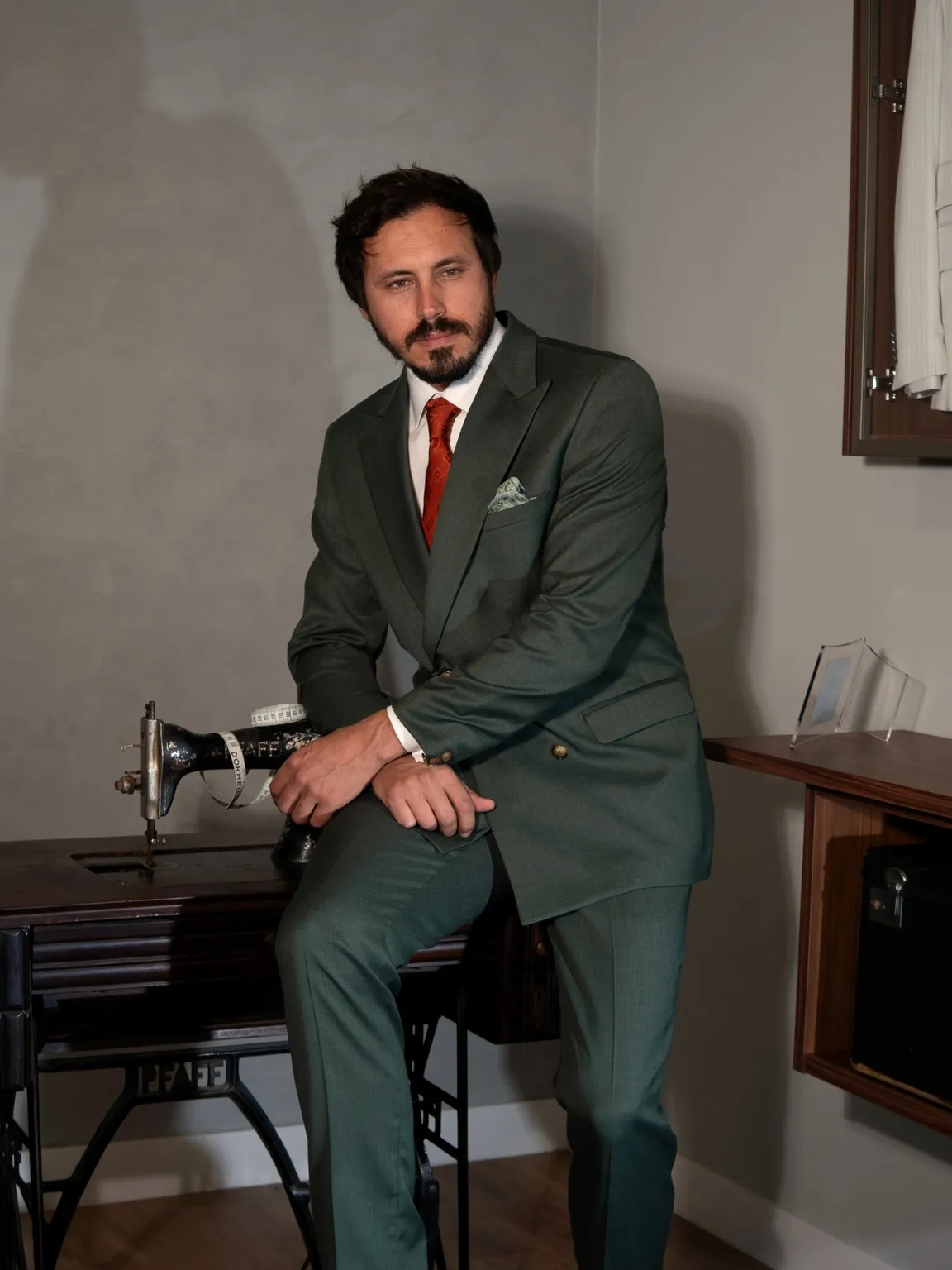 Terno verde com gravata vermelha, sentado na mesa de costura